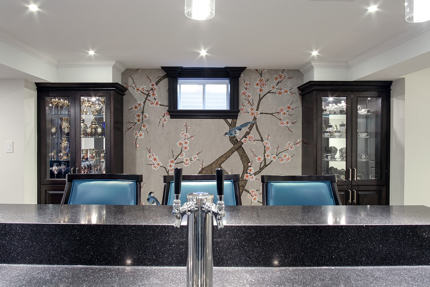 Home bar with patterned feature wall, lit glass shelving and blue leather stools — Burrowhill basement, Studio Kimi Mississauga